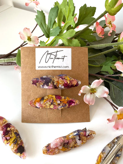 Close-up of handmade resin alligator hair clips with dried flowers and gold leaf on branded packaging card, surrounded by additional clips scattered on a white surface