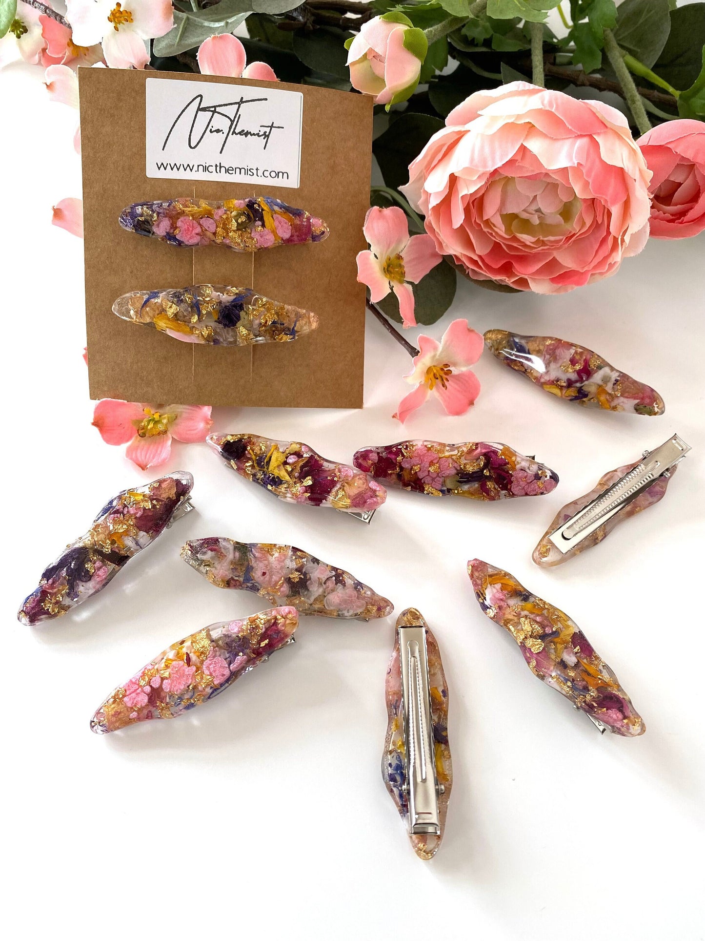 Two handmade resin alligator hair clips on branded packaging card in front of flowers, with additional clips scattered showing resin designs and silver alligator clip backs