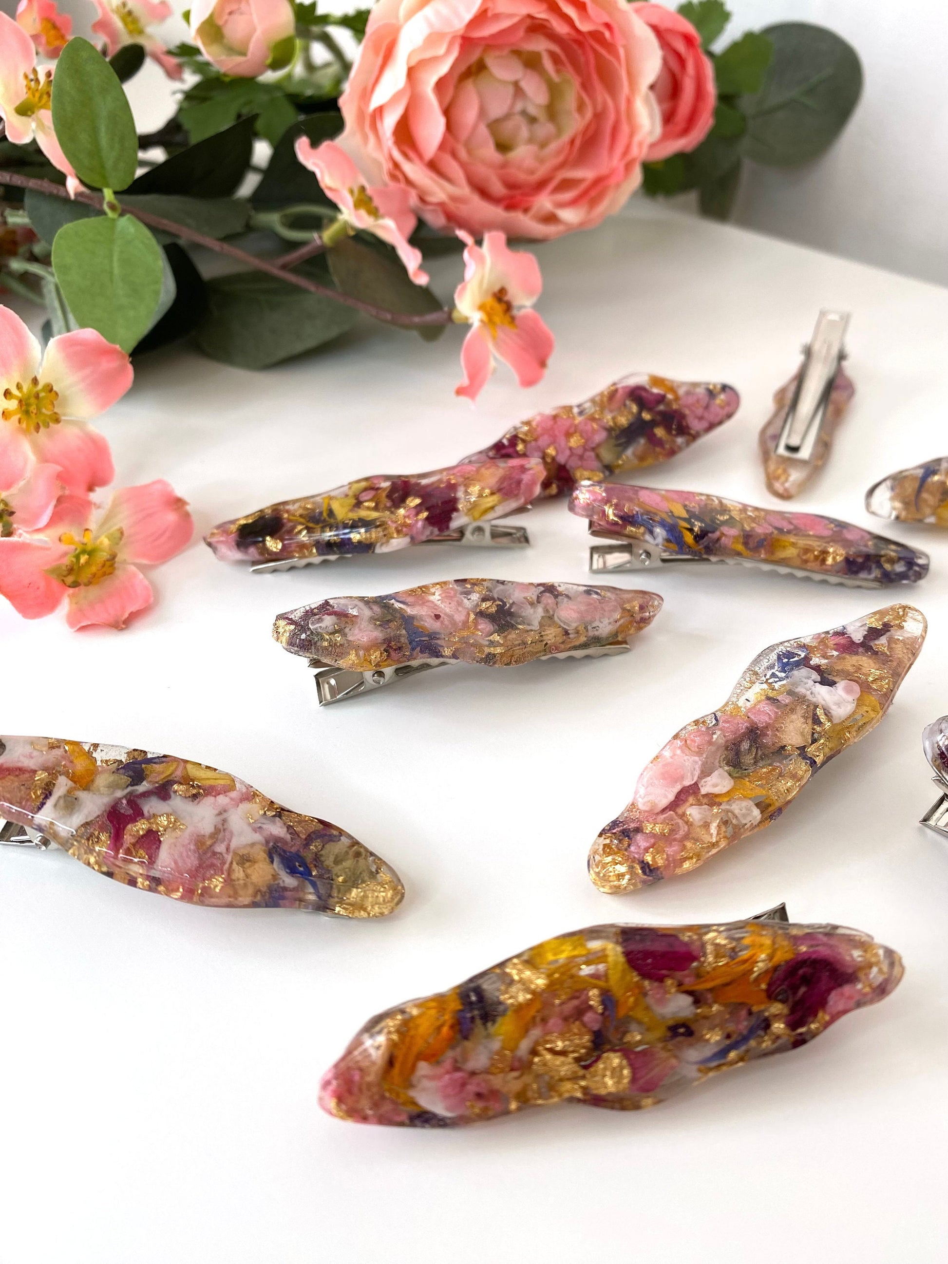 Handmade resin alligator hair clips with dried flowers and gold leaf scattered on a white surface, showing unique resin designs and silver clip backs