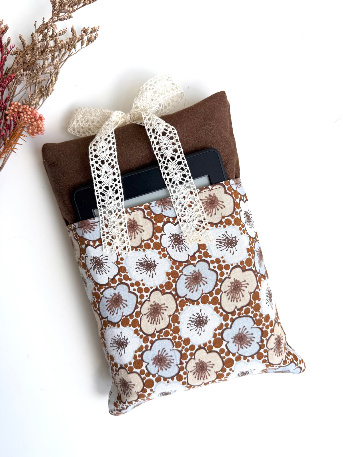 Brown faux suede book sleeve with a retro cream and pale blue floral front pocket holding a black e-reader, finished with a cream lace bow.