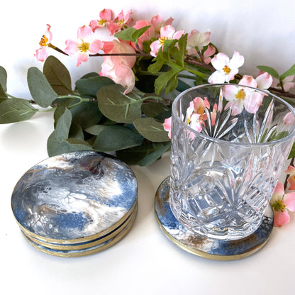 Set of 4 handmade round slate resin coasters with blue, white, black, and gold epoxy design and gold-painted edges, three stacked with the fourth next to them holding a water glass