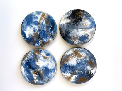 Top-down view of 4 handmade round slate resin coasters with blue, white, black, and gold epoxy designs and gold-painted edges – each coaster is unique and one-of-a-kind.