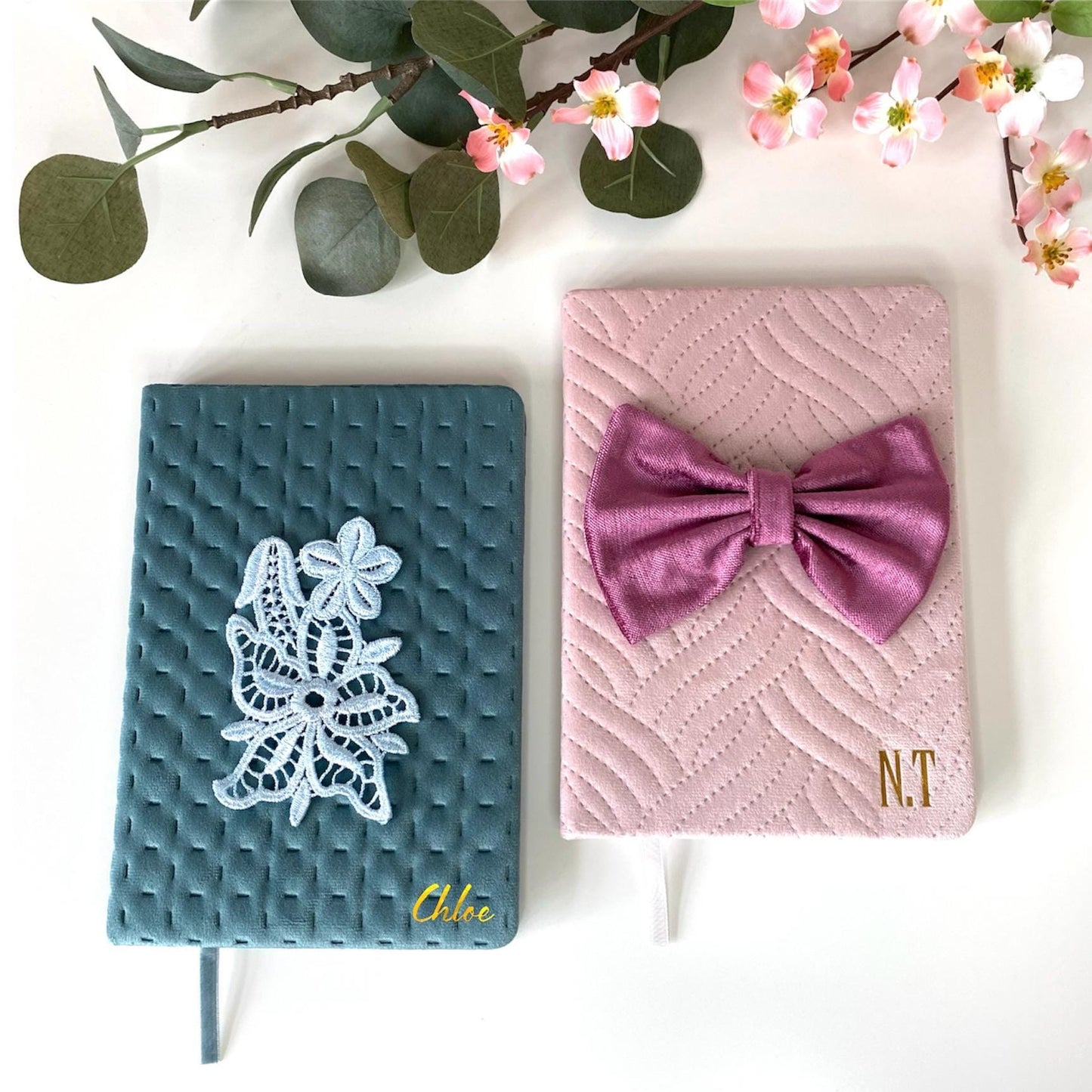 Top-down view of A5 luxury velvet notebooks on a white surface with flowers; teal notebook with floral appliqué personalised with a name and pink notebook with hand-sewn bow personalised with initials