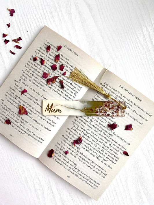 White and green resin bookmark with dried roses and gold leaf, personalised with gold vinyl, placed inside an open book, handmade bookmark for readers or gifts