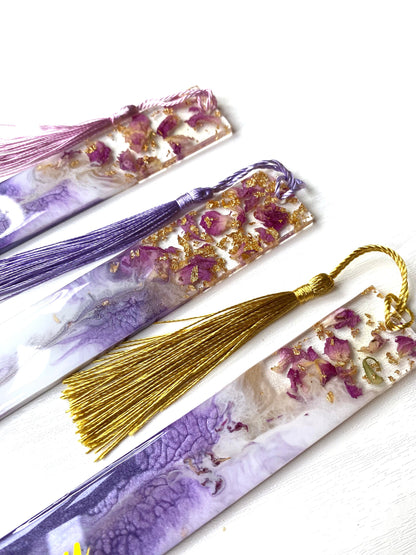 Close-up of three white and purple resin bookmarks with dried roses and gold leaf, one personalised on a purple base, one non-personalised on a white base, and one non-personalised on a purple base, showing handmade details, ideal bookmarks for readers or gifts.