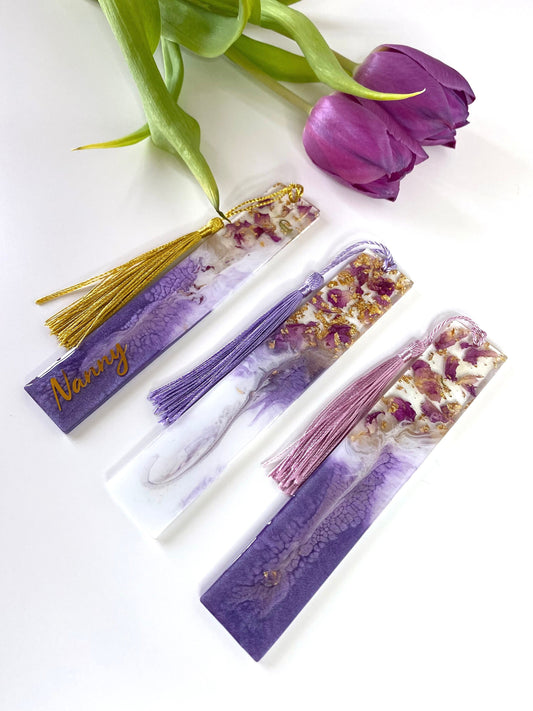 Three white and purple resin bookmarks with dried roses and gold leaf, one personalised with gold vinyl on a purple base, one non-personalised on a white base, and one non-personalised on a purple base, handmade bookmarks for readers or gifts.