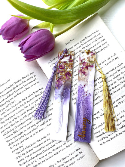 White and purple resin bookmarks with dried roses and gold leaf, both personalised with gold vinyl, placed inside an open book, handmade bookmarks for readers or gifts.